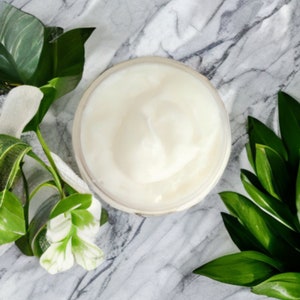 May include: A white cream in a clear jar on a marble surface with green leaves.