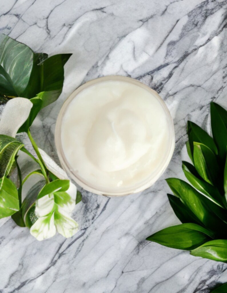 May include: A white cream in a clear jar on a marble surface with green leaves.