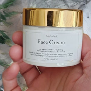 May include: A white jar with a gold lid labeled "Face Cream" with the text "All Natural/Oil Free/Hydrating W/Hyaluronic acid | Green Tea Extract" and a list of ingredients. The jar is 2.54 ounces (72g).