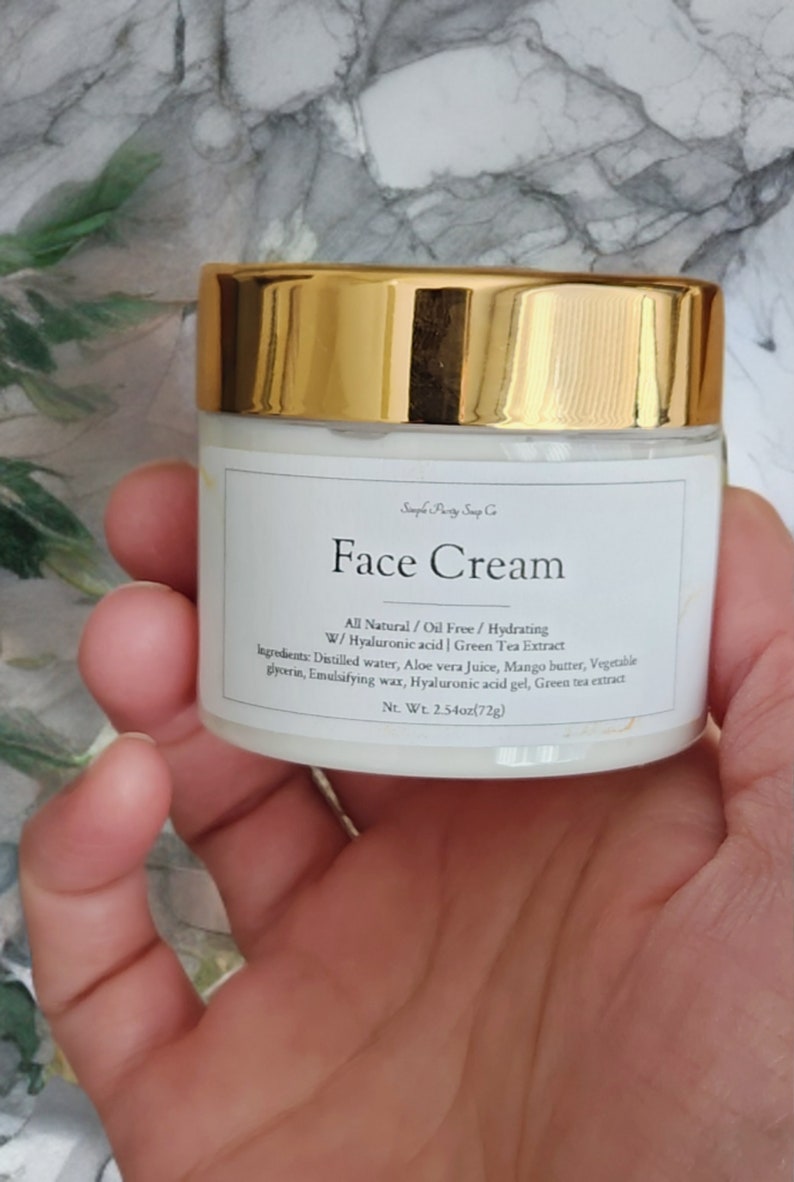May include: A white jar with a gold lid labeled "Face Cream" with the text "All Natural/Oil Free/Hydrating W/Hyaluronic acid | Green Tea Extract" and a list of ingredients. The jar is 2.54 ounces (72g).