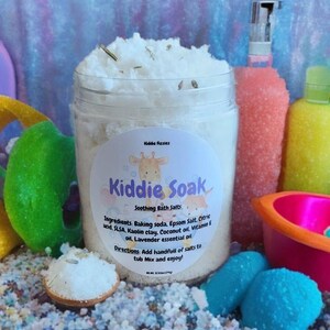 May include: A white jar of bath salts labeled "Kiddie Soak Soothing Bath Salts" with a cartoon giraffe on the label. The jar is surrounded by colorful bath toys and bath salts.