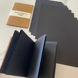 May include: An open black sketchbook with black pages, a closed brown notebook with a white band that reads "JustGee Paper Co. A5 Black", and a stack of black paper sheets. The pages are a deep black color.