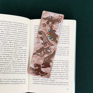 Könnte beinhalten: Ein skurriles Lesezeichen mit Schokoladen-Dinosaurier-Designs und Streuseln. Das Lesezeichen besteht aus klarem Acryl und befindet sich in einem Buch.