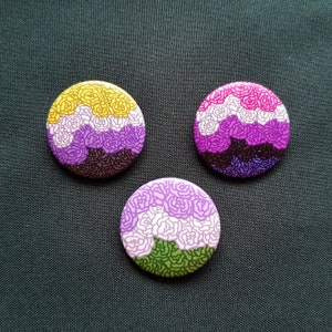 May include: Three round buttons with floral designs in various color schemes. One button features yellow, white, purple, and brown. Another has pink, white, purple, brown, and blue. The third button has purple, white, and green. All buttons are on a dark green surface.
