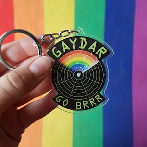 Könnte beinhalten: Ein klarer Acryl-Schlüsselanhänger mit einem silbernen Schlüsselring und einer Kette. Der Schlüsselanhänger zeigt ein schwarzes Radar-Design mit einem Regenbogenbogen und dem Text "GAYDAR GO BRRR" in Neongrün. Der Hintergrund ist eine Regenbogenflagge.