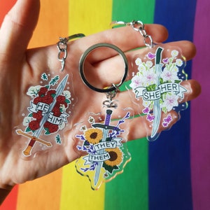 Könnte beinhalten: Drei transparente Acryl-Schlüsselanhänger mit Schwertmotiven. Einer zeigt rote Rosen und den Text "HE HIM", ein anderer Sonnenblumen und den Text "THEY THEM", und der letzte weiße Blumen und den Text "SHE HER". Jeder Schlüsselanhänger hat einen silbernen Schlüsselring.