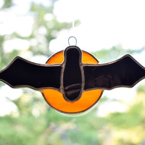 May include: A stained glass bat with black wings and a body, hanging in front of a bright orange circle.
