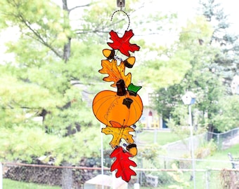 May include: A stained glass pumpkin with fall leaves and acorns hanging from a window. The pumpkin is orange with a green stem and brown vines. The leaves are red, orange, and yellow. The acorns are brown.