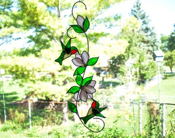 May include: A stained glass hummingbird and flower suncatcher with green leaves and white flowers. The suncatcher is hanging in a window with a blurry background of green trees and a fence.