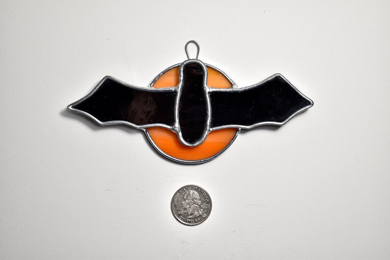 May include: A stained glass bat ornament with a black bat silhouette and an orange circle background. The ornament is approximately the size of a US quarter.