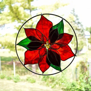 May include: A stained glass window hanging features a red poinsettia flower with black and green accents. The flower is in a circular frame.