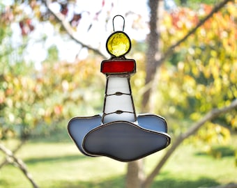 May include: A stained glass lighthouse ornament with a yellow light, red roof, and white tower. The lighthouse is on a blue cloud-shaped base.