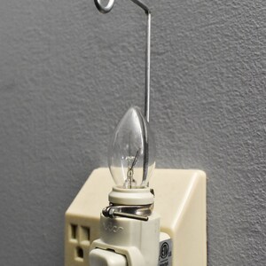 May include: A white night light with a clear light bulb and a metal hook. The night light has a switch and is plugged into an outlet. The outlet has a UL listed sticker.