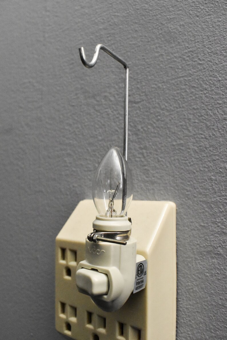 May include: A white night light with a clear light bulb and a metal hook. The night light has a switch and is plugged into an outlet. The outlet has a UL listed sticker.