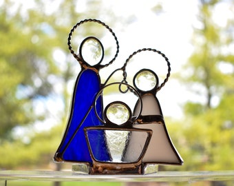 May include: A stained glass nativity scene with a blue Mary, white Joseph, and a baby Jesus in a manger. The figures are outlined in silver wire and have haloes.