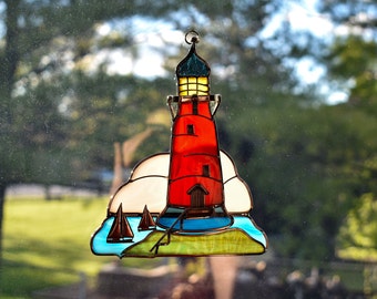 May include: A stained glass window hanging features a red lighthouse with a white cloud behind it. The lighthouse has a blue roof and yellow windows. There are three small sailboats in the water in front of the lighthouse.