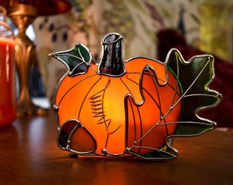 May include: A stained glass pumpkin candle holder with an orange pumpkin, green leaves, and a brown acorn. The candle holder is lit and glowing.