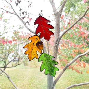 May include: A stained glass autumn leaf decoration featuring three oak leaves in red, yellow, and green, with a small acorn. The leaves are suspended from a wire, perfect for fall decor.