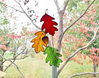 May include: A stained glass autumn leaf decoration featuring three oak leaves in red, yellow, and green, with a small acorn. The leaves are suspended from a wire, perfect for fall decor.