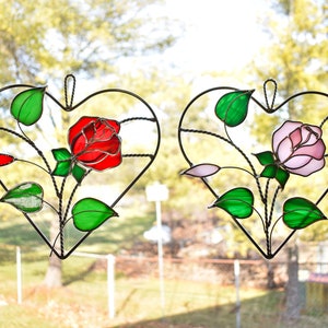 May include: Two heart-shaped stained glass panels with rose designs. One features a red rose, the other a pink rose, both with green leaves and a black metal frame. The panels are hanging in front of a blurred outdoor background.
