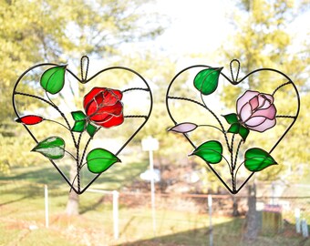 May include: Two heart-shaped stained glass panels with rose designs. One features a red rose, the other a pink rose, both with green leaves and a black metal frame. The panels are hanging in front of a blurred outdoor background.