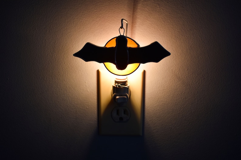May include: A nightlight shaped like a bat with a stained glass design. The nightlight is plugged into an electrical outlet and is lit up.