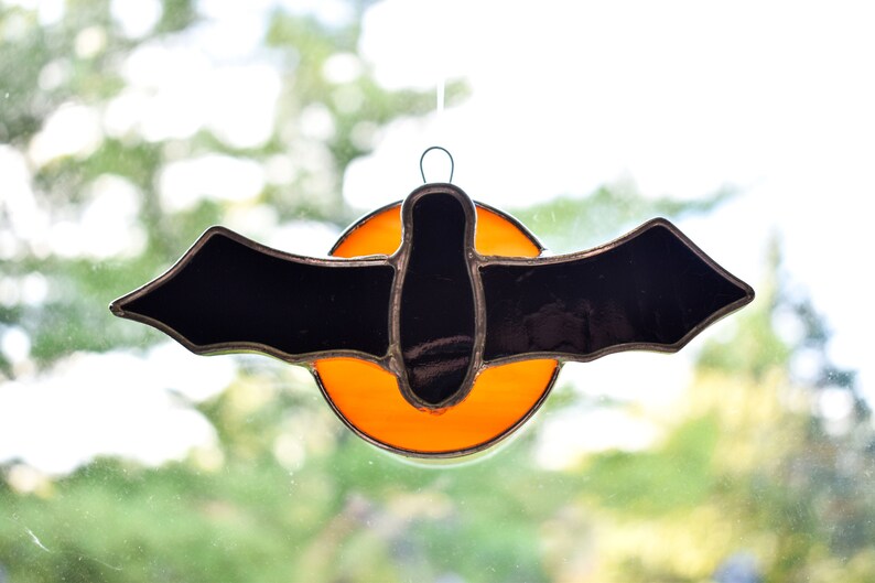 May include: A stained glass bat with black wings and a body, hanging in front of a bright orange circle.
