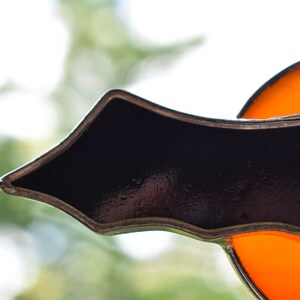 May include: A stained glass window decoration featuring a black bat with orange accents. The bat is made of glass and has a metal frame.