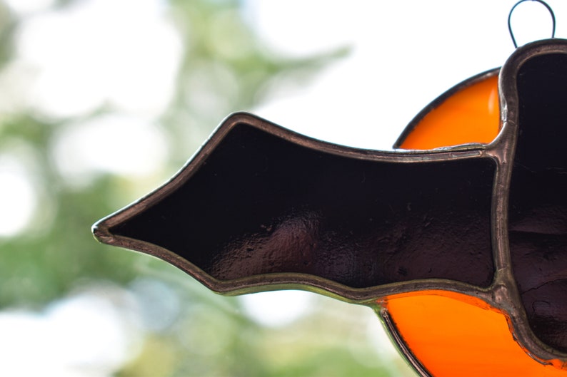 May include: A stained glass window decoration featuring a black bat with orange accents. The bat is made of glass and has a metal frame.