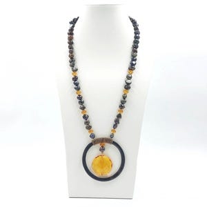 Puede incluir: Un collar largo con perlas de color gris oscuro de forma irregular intercaladas con pequeñas cuentas translúcidas de color ámbar. Un gran colgante de anillo negro cuelga del centro, sosteniendo un dije de vidrio redondo y facetado.