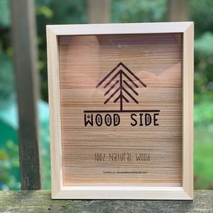 May include: A wooden frame with a light wood finish. The frame has a clear glass front and a brown wood back. The frame features a stylized tree design with the text "WOOD SIDE" and "100% Natural Wood". The text "Contact us: woodsideframe@gmail.com" is also included.