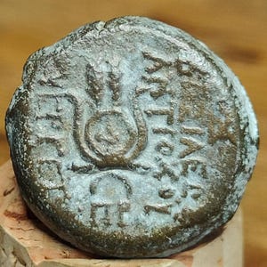 May include: A worn, bronze coin featuring embossed text and symbols. The coin has an aged appearance with an uneven shape. The design includes a wheat stalk and Greek lettering, suggesting an ancient origin. The coin is a weathered bronze colour.