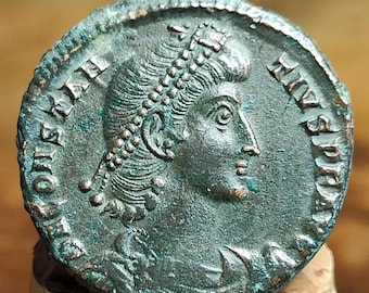 Op de afbeelding: Een verweerde, bronzen Romeinse munt. De munt toont een profiel van een persoon met een lauwerkrans en tekst rond de rand. De munt heeft een groene patina en is gemonteerd op een houten voet.