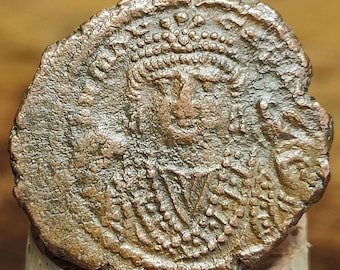 Könnte beinhalten: Eine verwitterte, bronzefarbene Münze mit einem detaillierten Porträt einer Person mit Krone. Die Oberfläche der Münze ist strukturiert und abgenutzt, mit sichtbaren Altersspuren. Das Porträt ist zentriert, mit komplizierten Details in Haar und Gesichtszügen.