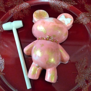 Może przedstawiać: Różowy i złoty miś pluszowy z białą kokardką na głowie. Miś siedzi na czerwonym talerzu z białym młotkiem obok.