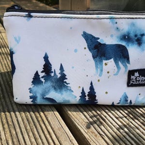 Könnte beinhalten: Weiße Reißverschlusstasche mit einem Aquarell-Design eines Wolfes, der zum Mond heult, umgeben von Bäumen und einem blau-weißen Sternenhimmel. Die Tasche hat ein kleines schwarzes Etikett mit dem Text "Dino Fashion".