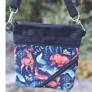 May include: A navy blue crossbody bag with a whimsical forest animal print. The bag features a black top section, a diagonal zip, and a black adjustable strap. The print includes deer, bears, rabbits, and foxes in shades of red, blue, and white.