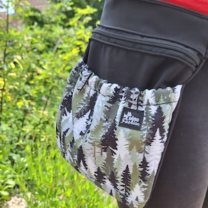 Könnte beinhalten: Ein schwarzer Hüftgürtel mit einer großen Reißverschlusstasche an der Seite. Die Tasche ist mit einem Stoff mit Waldmotiv und dem Text "pino fashion" gefüttert.