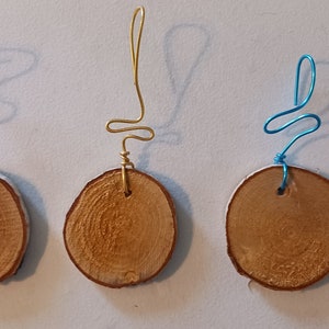 May include: Three wooden ornaments with wire hangers. The ornaments are round and have a natural wood finish. The hangers are different colors: purple, gold, and blue.