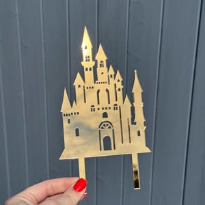 May include: A gold-coloured castle cake topper with multiple turrets, windows, and a central arched doorway. The topper is held up by two rectangular posts. The background is a grey wooden surface.