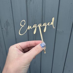 May include: A gold-coloured cake topper with the word "Engaged" in a cursive font. The topper is held by a thin stem, perfect for inserting into a cake. The background is a weathered grey wooden surface.