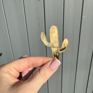 K&ouml;nnte beinhalten: Ein goldfarbenes, kaktusf&ouml;rmiges Dekorationsobjekt. Der Kaktus hat ein detailliertes Design mit kleinen Vertiefungen. Der Artikel wird vor einem grauen Hintergrund gehalten. Der Kaktus ist ein beliebtes Dekorationsst&uuml;ck im W&uuml;stenstil.