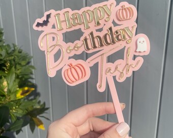 Może przedstawiać: Różowo-złota dekoracja na tort z motywem Halloween z napisem "Happy Birthday Tash" i "Boo" w złotych literach. Ozdoba zawiera nietoperza, dynie i ducha. Ozdoba jest trzymana przez różowy patyczek.