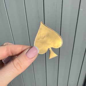 May include: A gold-coloured spade-shaped object. The object is held against a grey wooden background. The spade shape is smooth and reflective, with a glossy finish. The edges of the spade are clearly defined.