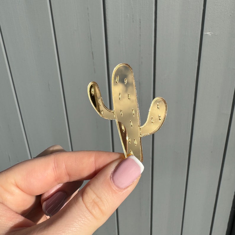 K&ouml;nnte beinhalten: Ein goldfarbenes, kaktusf&ouml;rmiges Dekorationsobjekt. Der Kaktus hat eine glatte, reflektierende Oberfl&auml;che und wird vor einem grauen Hintergrund gehalten. Der Kaktus hat zwei Seitenarme und kleine, gleichm&auml;&szlig;ig verteilte Punkte.