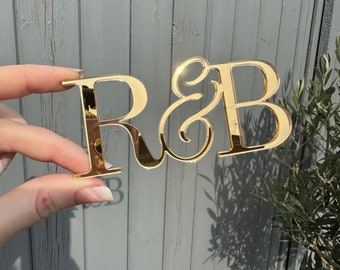 May include: A gold-coloured, mirrored acrylic sign displaying the letters "R&B" in a decorative font. The sign is held up against a grey wooden background, with sunlight reflecting off the shiny surface. The letters are approximately 10 cm wide.