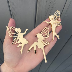 May include: Three gold-coloured fairy cutouts with detailed wing designs. One fairy is standing, another is seated, and the third is in a dancing pose. The cutouts are made of a shiny, reflective material, and are likely for decorative purposes.