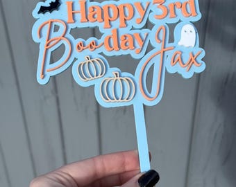 May include: A light blue cake topper with orange text reading "Happy 3rd Boo-day Jax". It features a black bat, a white ghost, and two orange pumpkins. The topper is held by a hand.