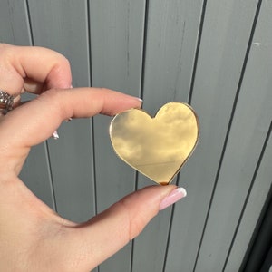 May include: A gold-coloured heart-shaped object. The heart is smooth and reflective, with the sky reflected in its surface. The object is held between fingers against a grey background.