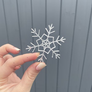 May include: A clear, snowflake-shaped decoration with detailed geometric cutouts. The snowflake is held between fingers against a grey background. The design features six symmetrical arms with delicate, branch-like details at the tips, ideal for winter decor.